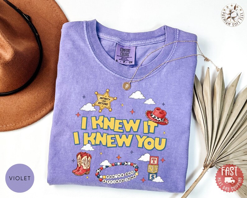 May include: A violet short-sleeved t-shirt featuring a graphic print with the text I KNEW IT I KNEW YOU, a yellow sheriff star, a red cowboy hat, red cowboy boots, a beaded bracelet with the text YOU'VE GOT A FRIEND IN ME, and alphabet blocks.