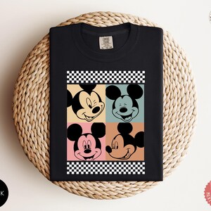 Peut inclure: T-shirt noir avec un motif de style pop art du visage de Mickey Mouse dans quatre blocs de couleurs diff&eacute;rents. Le motif comprend une bordure &agrave; carreaux noirs et blancs. Le t-shirt est pli&eacute; sur une surface tiss&eacute;e.