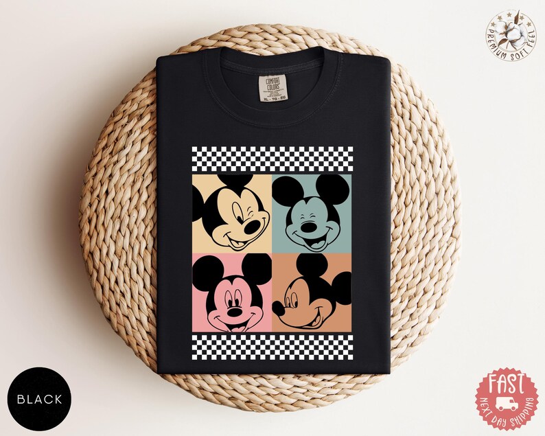 Peut inclure: T-shirt noir avec un motif de style pop art du visage de Mickey Mouse dans quatre blocs de couleurs diff&eacute;rents. Le motif comprend une bordure &agrave; carreaux noirs et blancs. Le t-shirt est pli&eacute; sur une surface tiss&eacute;e.