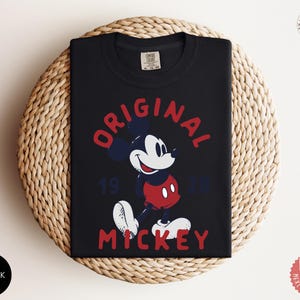 May include: Black t-shirt featuring a graphic of Mickey Mouse in red, white, and blue. The words "ORIGINAL MICKEY 1928" are printed above and below the character. The shirt is folded on a woven, circular surface.
