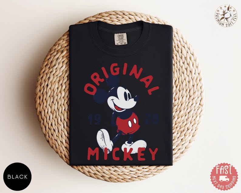 May include: Black t-shirt featuring a graphic of Mickey Mouse in red, white, and blue. The words "ORIGINAL MICKEY 1928" are printed above and below the character. The shirt is folded on a woven, circular surface.