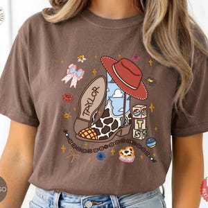May include: A brown short-sleeve t-shirt featuring a graphic print of a cowboy boot with a cow print pattern, a red cowboy hat, a blue sky background, the name TAYLOR, the number 13, stacked blocks with letters, and a beaded bracelet design.
