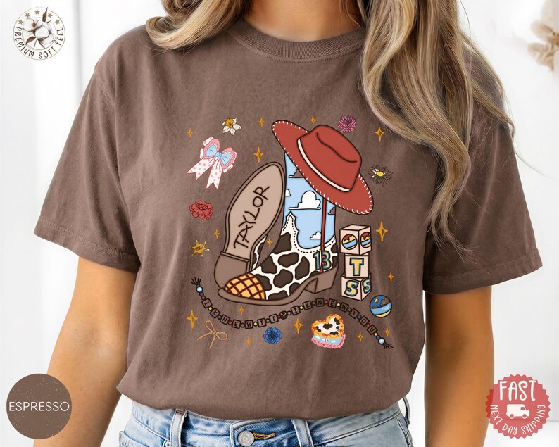 May include: A brown short-sleeve t-shirt featuring a graphic print of a cowboy boot with a cow print pattern, a red cowboy hat, a blue sky background, the name TAYLOR, the number 13, stacked blocks with letters, and a beaded bracelet design.