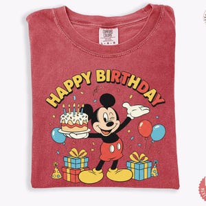 K&ouml;nnte beinhalten: Rotes T-Shirt mit einem Mickey Mouse-Geburtstagsmotiv. Das Design umfasst einen Kuchen, Luftballons und Geschenkverpackungen mit dem Schriftzug "Happy Birthday". Das T-Shirt ist gefaltet.