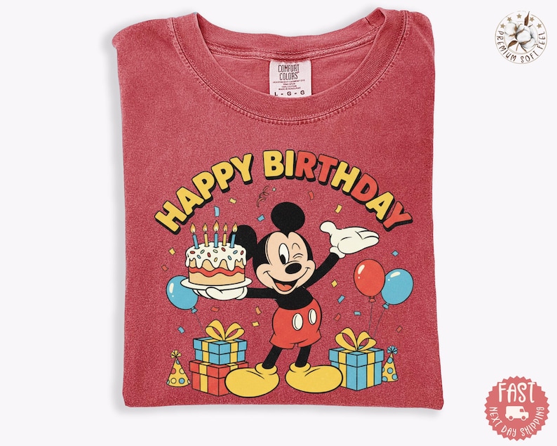 K&ouml;nnte beinhalten: Rotes T-Shirt mit einem Mickey Mouse-Geburtstagsmotiv. Das Design umfasst einen Kuchen, Luftballons und Geschenkverpackungen mit dem Schriftzug "Happy Birthday". Das T-Shirt ist gefaltet.