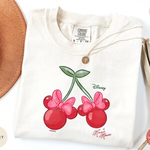 Peut inclure: T-shirt ivoire avec un motif de cerises Minnie Mouse. Les cerises sont rouges avec des n&oelig;uds roses et des feuilles vertes, avec le logo Disney. Le t-shirt a un col rond et des manches courtes. Les mots "Minnie Mouse" sont &eacute;crits en cursive.