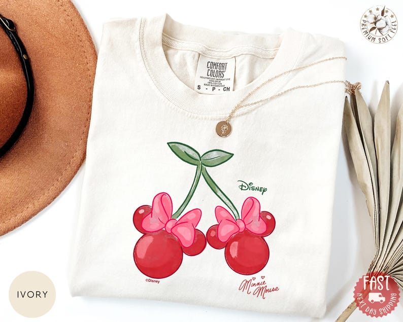 Peut inclure: T-shirt ivoire avec un motif de cerises Minnie Mouse. Les cerises sont rouges avec des n&oelig;uds roses et des feuilles vertes, avec le logo Disney. Le t-shirt a un col rond et des manches courtes. Les mots "Minnie Mouse" sont &eacute;crits en cursive.