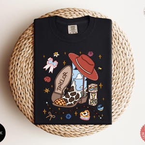 May include: A black t-shirt featuring a graphic print of a cowboy boot with a cow print pattern, a red hat, and the name Taylor. The design includes small blocks with the letters T, S, and a rainbow, along with stars, flowers, and a bow.