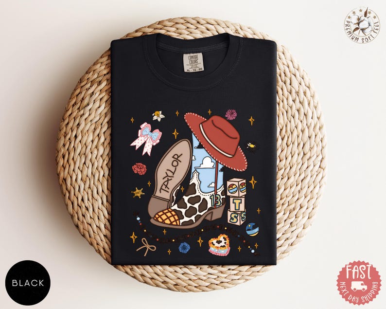 May include: A black t-shirt featuring a graphic print of a cowboy boot with a cow print pattern, a red hat, and the name Taylor. The design includes small blocks with the letters T, S, and a rainbow, along with stars, flowers, and a bow.