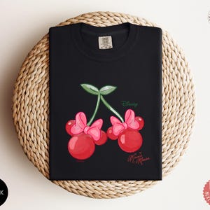 Peut inclure: T-shirt noir avec un motif fantaisiste de deux cerises rouges avec des n&oelig;uds roses, des feuilles vertes et le texte "Disney Minnie Mouse". Le t-shirt est pli&eacute; sur une surface tiss&eacute;e.