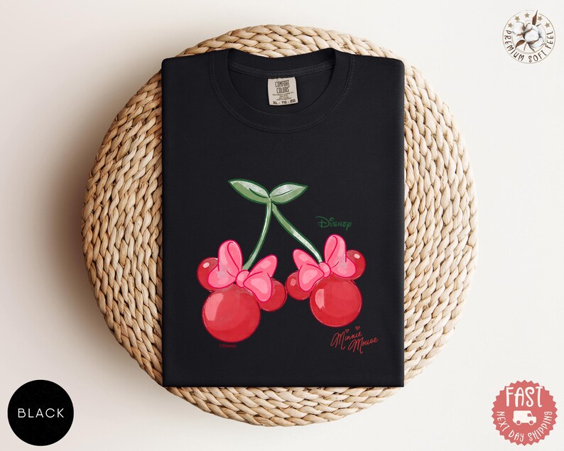 Peut inclure: T-shirt noir avec un motif fantaisiste de deux cerises rouges avec des n&oelig;uds roses, des feuilles vertes et le texte "Disney Minnie Mouse". Le t-shirt est pli&eacute; sur une surface tiss&eacute;e.