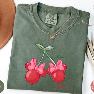 Peut inclure: T-shirt vert mousse avec un motif de cerises avec des n&oelig;uds Minnie Mouse. Le motif comprend le mot "Disney". Le t-shirt a un col rond et des manches courtes. Un collier en or est drap&eacute; sur le t-shirt.
