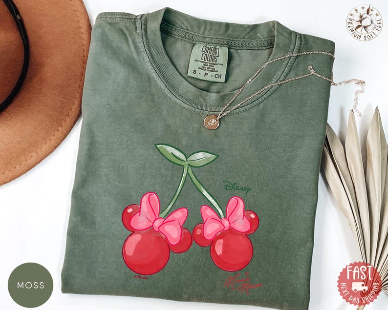 Peut inclure: T-shirt vert mousse avec un motif de cerises avec des n&oelig;uds Minnie Mouse. Le motif comprend le mot "Disney". Le t-shirt a un col rond et des manches courtes. Un collier en or est drap&eacute; sur le t-shirt.
