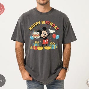 K&ouml;nnte beinhalten: Dunkelgraues T-Shirt mit einer Grafik von Mickey Mouse, die eine Geburtstagstorte h&auml;lt. Der Text "HAPPY BIRTHDAY" befindet sich &uuml;ber der Figur. Ballons und Geschenke erg&auml;nzen das Design.