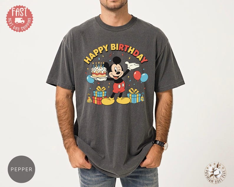 K&ouml;nnte beinhalten: Dunkelgraues T-Shirt mit einer Grafik von Mickey Mouse, die eine Geburtstagstorte h&auml;lt. Der Text "HAPPY BIRTHDAY" befindet sich &uuml;ber der Figur. Ballons und Geschenke erg&auml;nzen das Design.