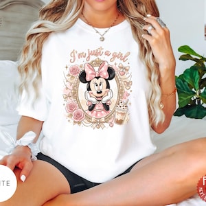 May include: A white t-shirt featuring a graphic print of Minnie Mouse inside an ornate gold frame, surrounded by pink roses and bows. The text "I'm just a girl" is printed above the character, who is holding a beverage in a clear plastic cup.