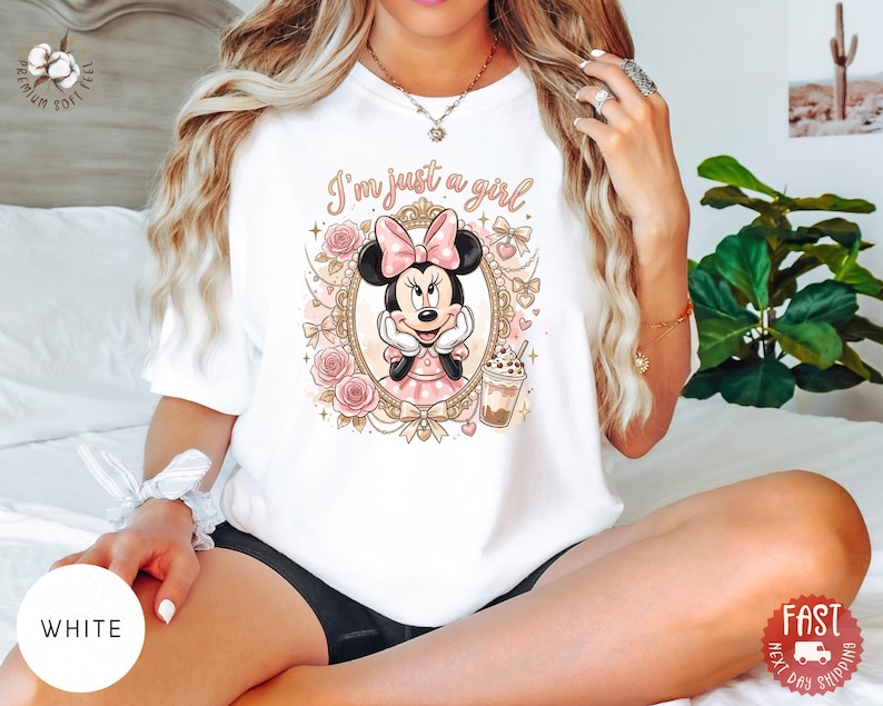 May include: A white t-shirt featuring a graphic print of Minnie Mouse inside an ornate gold frame, surrounded by pink roses and bows. The text "I'm just a girl" is printed above the character, who is holding a beverage in a clear plastic cup.