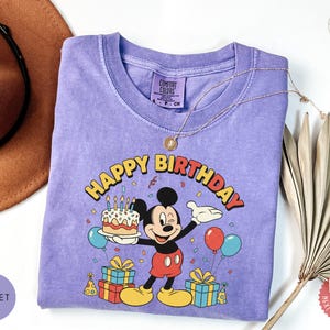 K&ouml;nnte beinhalten: Violettes T-Shirt mit einem Aufdruck von Mickey Mouse, der eine Geburtstagstorte h&auml;lt, mit den Worten "Happy Birthday" dar&uuml;ber. Das Design umfasst Luftballons, Konfetti und verpackte Geschenke. Das Shirt hat einen Rundhalsausschnitt und kurze &Auml;rmel.