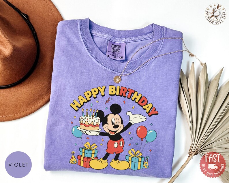 K&ouml;nnte beinhalten: Violettes T-Shirt mit einem Aufdruck von Mickey Mouse, der eine Geburtstagstorte h&auml;lt, mit den Worten "Happy Birthday" dar&uuml;ber. Das Design umfasst Luftballons, Konfetti und verpackte Geschenke. Das Shirt hat einen Rundhalsausschnitt und kurze &Auml;rmel.