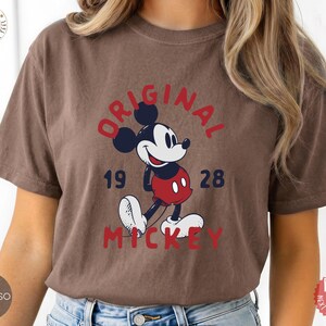 May include: Espresso-colored t-shirt featuring a classic Mickey Mouse graphic. The design includes the words "ORIGINAL MICKEY 1928" in red and navy blue. The shirt has short sleeves and a relaxed fit.