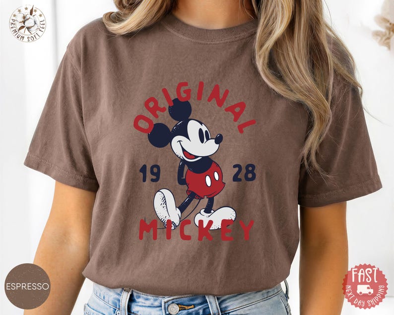 May include: Espresso-colored t-shirt featuring a classic Mickey Mouse graphic. The design includes the words "ORIGINAL MICKEY 1928" in red and navy blue. The shirt has short sleeves and a relaxed fit.