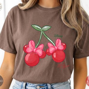 Peut inclure: T-shirt couleur expresso avec un motif de cerises de dessin anim&eacute; avec des n&oelig;uds inspir&eacute;s de Minnie Mouse. Les cerises sont rouges avec des n&oelig;uds roses et des tiges vertes. Le logo Disney est visible. C'est un v&ecirc;tement d&eacute;contract&eacute;.