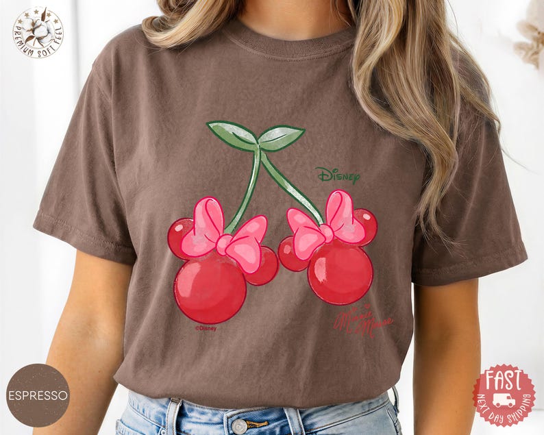 Peut inclure: T-shirt couleur expresso avec un motif de cerises de dessin anim&eacute; avec des n&oelig;uds inspir&eacute;s de Minnie Mouse. Les cerises sont rouges avec des n&oelig;uds roses et des tiges vertes. Le logo Disney est visible. C'est un v&ecirc;tement d&eacute;contract&eacute;.