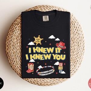 May include: A black t-shirt featuring a graphic print with the text I KNEW IT I KNEW YOU. The design includes a yellow sheriff star badge with the text INFINITY AND BEYOND, a red cowboy hat, red boots, a beaded bracelet with the text YOU'VE GOT A FRIEND IN ME, and a stack of three alphabet blocks.