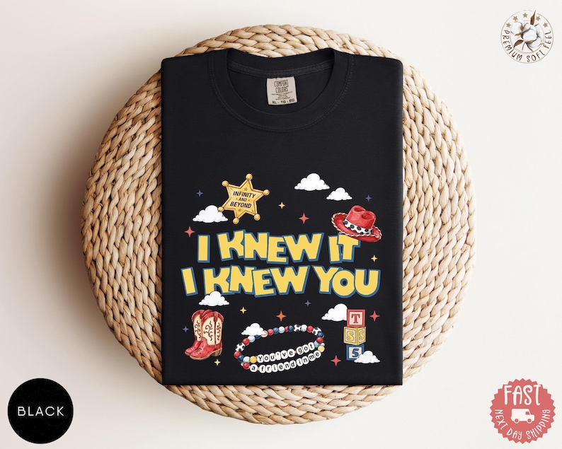 May include: A black t-shirt featuring a graphic print with the text I KNEW IT I KNEW YOU. The design includes a yellow sheriff star badge with the text INFINITY AND BEYOND, a red cowboy hat, red boots, a beaded bracelet with the text YOU'VE GOT A FRIEND IN ME, and a stack of three alphabet blocks.