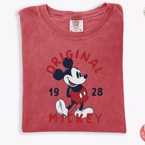 May include: A red t-shirt featuring a graphic of Mickey Mouse. The design includes the word "ORIGINAL" in a semi-circle above the character, with "1928" and "MICKEY" below. The shirt is folded and has a tag that says "COMFORT COLORS".