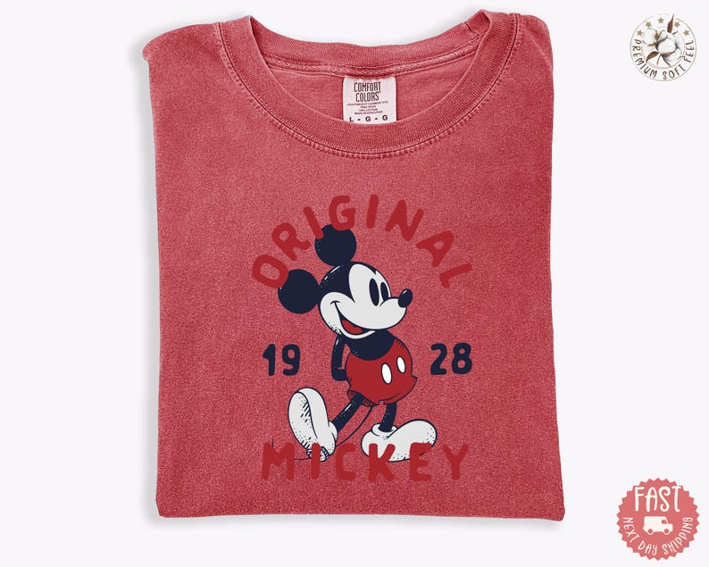 May include: A red t-shirt featuring a graphic of Mickey Mouse. The design includes the word "ORIGINAL" in a semi-circle above the character, with "1928" and "MICKEY" below. The shirt is folded and has a tag that says "COMFORT COLORS".