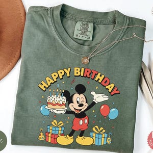 K&ouml;nnte beinhalten: Moosgr&uuml;nes T-Shirt mit einem Mickey Mouse-Geburtstagsmotiv. Das Design beinhaltet den Text "HAPPY BIRTHDAY", einen Kuchen, Luftballons und Geschenke. Das T-Shirt hat einen Rundhalsausschnitt und kurze &Auml;rmel.