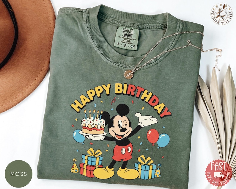 K&ouml;nnte beinhalten: Moosgr&uuml;nes T-Shirt mit einem Mickey Mouse-Geburtstagsmotiv. Das Design beinhaltet den Text "HAPPY BIRTHDAY", einen Kuchen, Luftballons und Geschenke. Das T-Shirt hat einen Rundhalsausschnitt und kurze &Auml;rmel.
