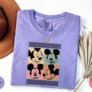 Peut inclure: Un t-shirt violet Comfort Colors avec un motif &agrave; quatre panneaux de visages de Mickey Mouse. Le design comprend des bordures &agrave; carreaux noirs et blancs et des arri&egrave;re-plans color&eacute;s. Un collier en or repose sur le t-shirt.