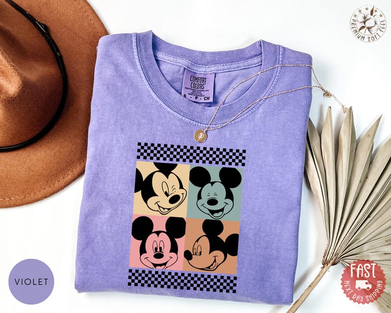 Peut inclure: Un t-shirt violet Comfort Colors avec un motif &agrave; quatre panneaux de visages de Mickey Mouse. Le design comprend des bordures &agrave; carreaux noirs et blancs et des arri&egrave;re-plans color&eacute;s. Un collier en or repose sur le t-shirt.