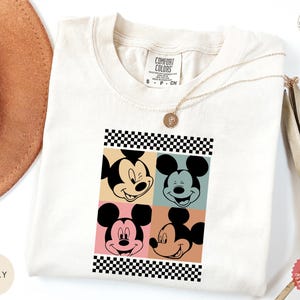 Peut inclure: T-shirt ivoire avec un motif de style pop art du visage de Mickey Mouse en quatre blocs de couleurs diff&eacute;rents. Le motif est encadr&eacute; d'un motif &agrave; carreaux noir et blanc. Col rond et manches courtes.