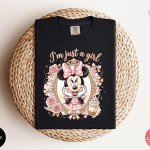 May include: A black t-shirt featuring a graphic of Minnie Mouse inside an ornate gold frame, surrounded by pink roses and bows. The text "I'm just a girl" is printed in a light pink script above the frame, with a small iced coffee drink nearby.