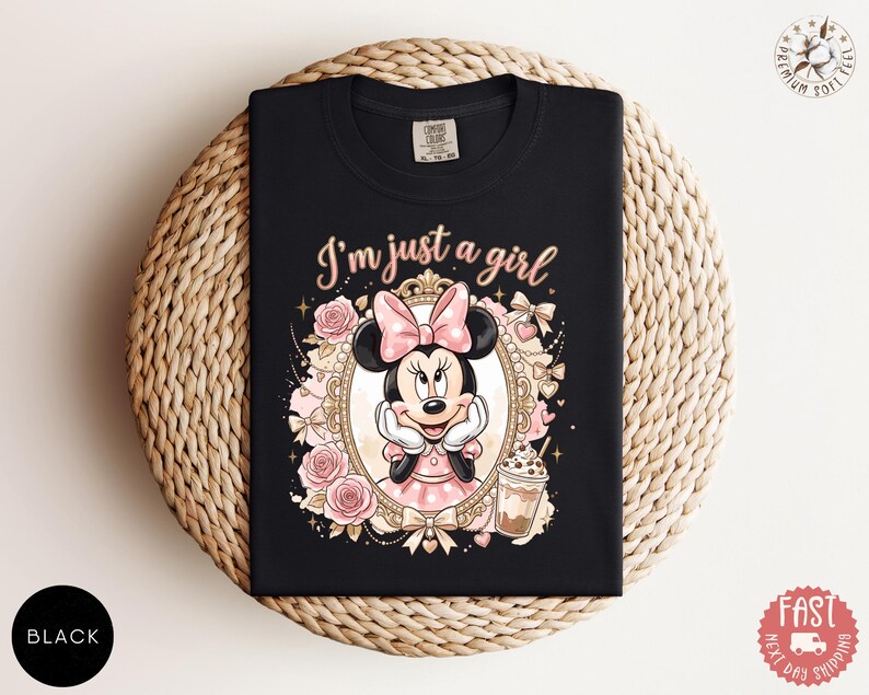 May include: A black t-shirt featuring a graphic of Minnie Mouse inside an ornate gold frame, surrounded by pink roses and bows. The text "I'm just a girl" is printed in a light pink script above the frame, with a small iced coffee drink nearby.