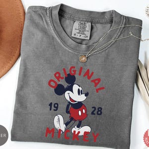 May include: A gray t-shirt featuring a graphic of Mickey Mouse in red, black, white, and blue. The text "ORIGINAL MICKEY 1928" is printed in red and blue. A silver necklace with a pendant is draped on the shirt.