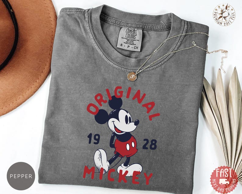 May include: A gray t-shirt featuring a graphic of Mickey Mouse in red, black, white, and blue. The text "ORIGINAL MICKEY 1928" is printed in red and blue. A silver necklace with a pendant is draped on the shirt.
