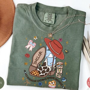 May include: A moss green short-sleeve t-shirt featuring a graphic print of a cowboy boot with a cow print pattern, a red hat, the name TAYLOR, the number 13, and stacked blocks with the letters B, T, and S, surrounded by small floral and star icons.
