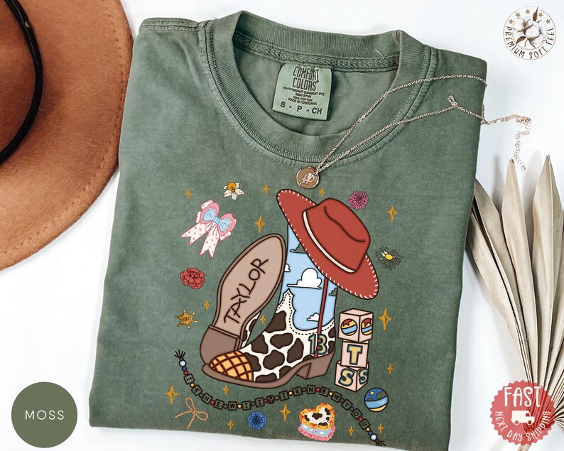 May include: A moss green short-sleeve t-shirt featuring a graphic print of a cowboy boot with a cow print pattern, a red hat, the name TAYLOR, the number 13, and stacked blocks with the letters B, T, and S, surrounded by small floral and star icons.