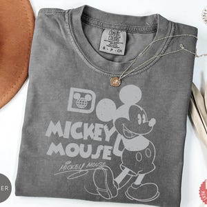 May include: A grey Comfort Colors t-shirt with a Mickey Mouse graphic and the words "Mickey Mouse". The shirt has a crew neck and short sleeves. A gold necklace is draped across the shirt.