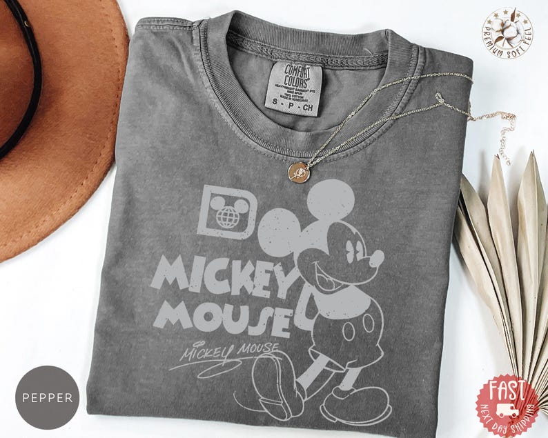 May include: A grey Comfort Colors t-shirt with a Mickey Mouse graphic and the words "Mickey Mouse". The shirt has a crew neck and short sleeves. A gold necklace is draped across the shirt.