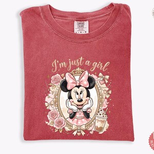 May include: A red short-sleeved t-shirt featuring a graphic of Minnie Mouse inside an ornate gold frame, surrounded by pink roses and bows. The text "I'm just a girl" is printed above the frame, and a small iced coffee drink is shown below.