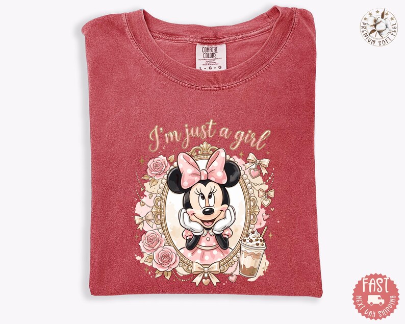 May include: A red short-sleeved t-shirt featuring a graphic of Minnie Mouse inside an ornate gold frame, surrounded by pink roses and bows. The text "I'm just a girl" is printed above the frame, and a small iced coffee drink is shown below.