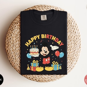 K&ouml;nnte beinhalten: Schwarzes T-Shirt mit einer farbenfrohen Grafik von Mickey Mouse, einer Geburtstagstorte, Luftballons und dem Schriftzug "HAPPY BIRTHDAY". Das Design umfasst Geschenkboxen und Konfetti und schafft ein festliches Geburtstagsthema.