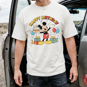 K&ouml;nnte beinhalten: Cremefarbenes T-Shirt mit einer bunten Grafik von Micky Maus, die eine Geburtstagstorte, Luftballons und Geschenke h&auml;lt. Die Worte "HAPPY BIRTHDAY" sind &uuml;ber Micky gebogen. Das Shirt hat eine lockere Passform.