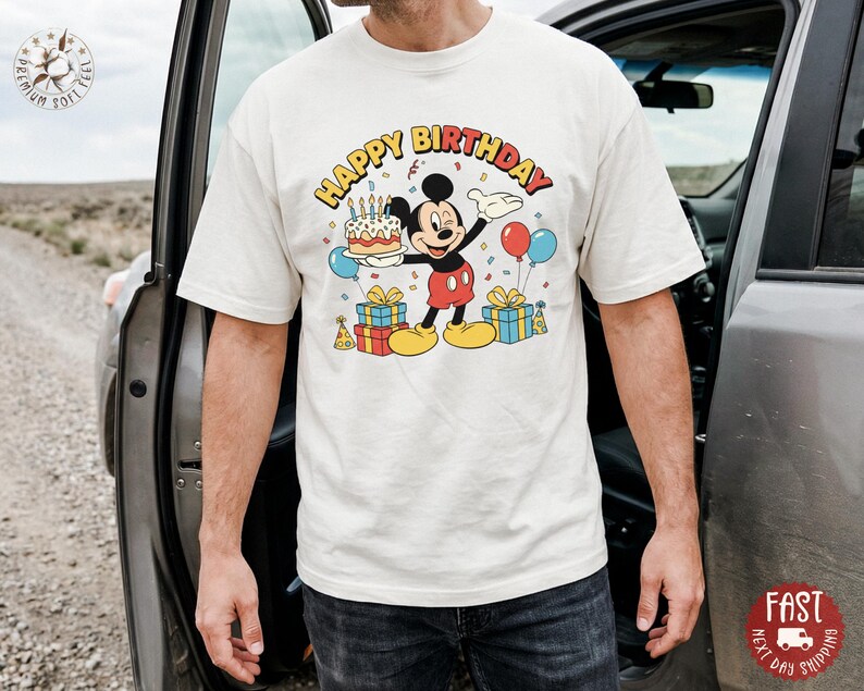 K&ouml;nnte beinhalten: Cremefarbenes T-Shirt mit einer bunten Grafik von Micky Maus, die eine Geburtstagstorte, Luftballons und Geschenke h&auml;lt. Die Worte "HAPPY BIRTHDAY" sind &uuml;ber Micky gebogen. Das Shirt hat eine lockere Passform.