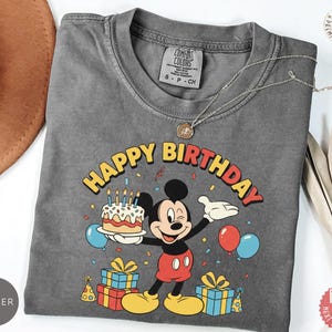 K&ouml;nnte beinhalten: Ein graues Comfort Colors T-Shirt mit einer bunten Grafik von Mickey Mouse, die eine Geburtstagstorte, Luftballons und Geschenke h&auml;lt. Die Worte "Happy Birthday" sind in Gelb und Rot gehalten. Eine silberne Halskette liegt &uuml;ber dem Shirt.